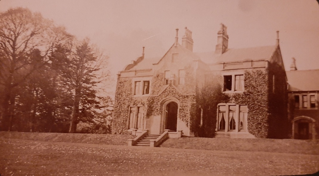 Sepia coloured photograph of the front of Annaghmakerrig House, about 1900. The house is covered in ivy, and has twelve windows and an arched front door, with a decorative roof, large chimney pots. The grass to the front is well manicured, and there are a group of evergreen and deciduous trees to the left of the house.